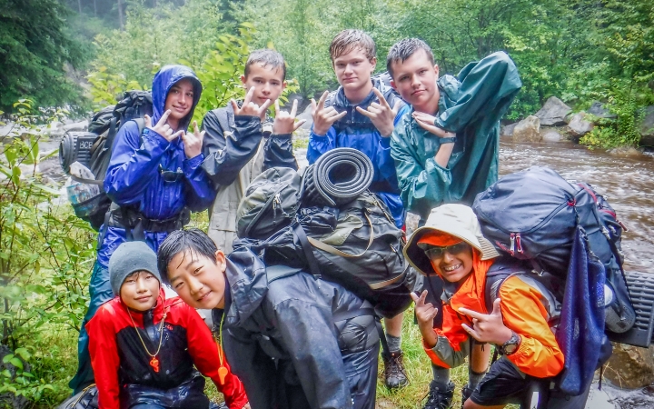 A group of young people wearing backpacks and rain gear pose for a photo beside a river in a densely wooded area. 